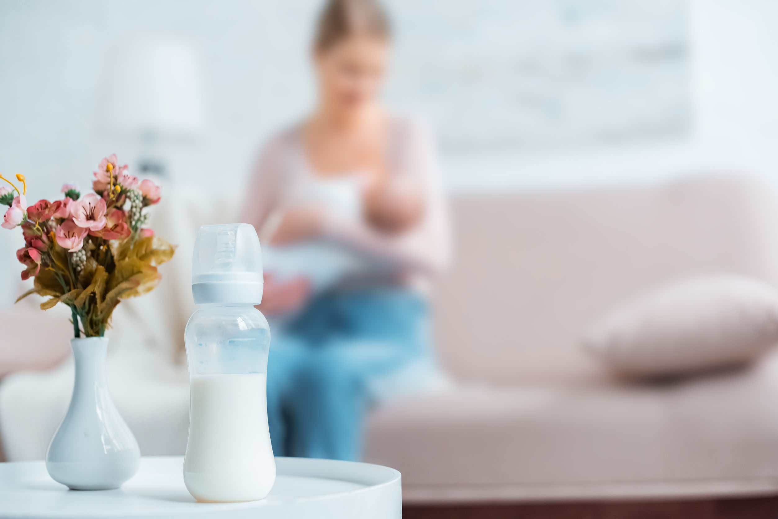 Photo of a bottle of breast milk. A mother is breastfeeding her child in the background.