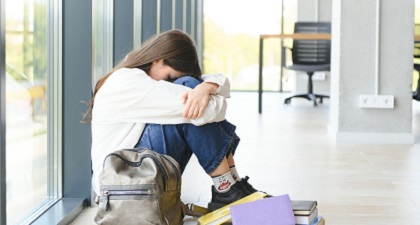 An upset teen girl sits on floor sadly with school bag and books on her side