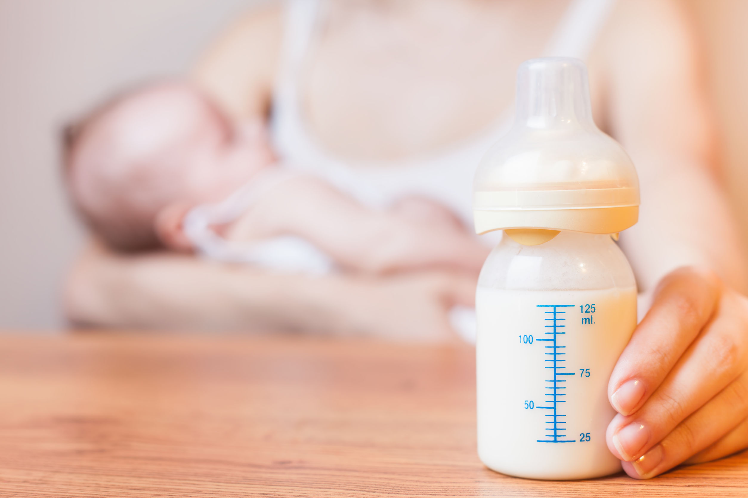 Photo of a bottle of breast milk. In the background: a woman is breastfeeding a baby.
