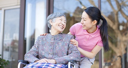 A woman takes care of an old lady who sits on a wheel chair. They smile to each other.