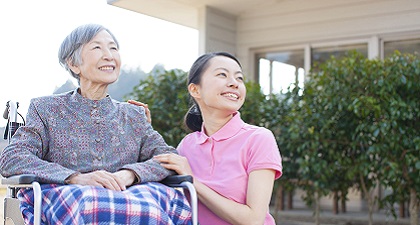 A carer is standing by the side of an old woman with a smiling face on a wheelchair