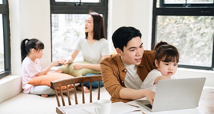 In a home setting, a mother is playing with her daughter on a sofa and a father is teaching his daughter with a laptop computer.